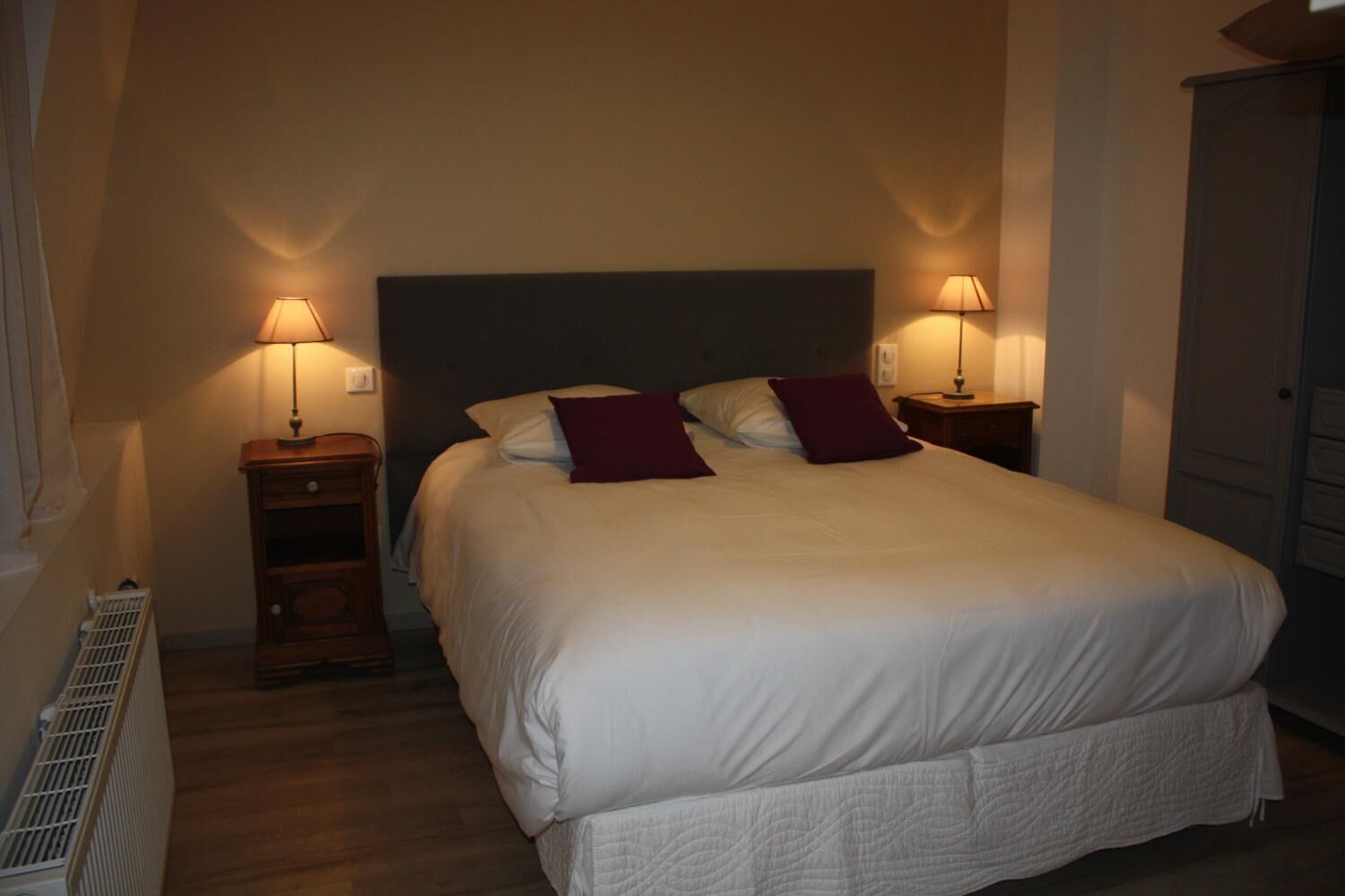Una cama bien tendida con ropa blanca y dos almohadas rojas, con dos lámparas de noche de madera a cada lado.