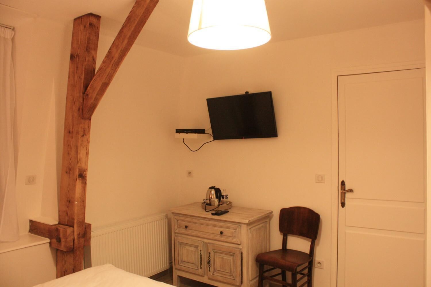 A small room with a wooden ceiling beam, a white radiator, a wooden dresser with a chair, a flat-screen TV mounted on the wall, and a closed door.