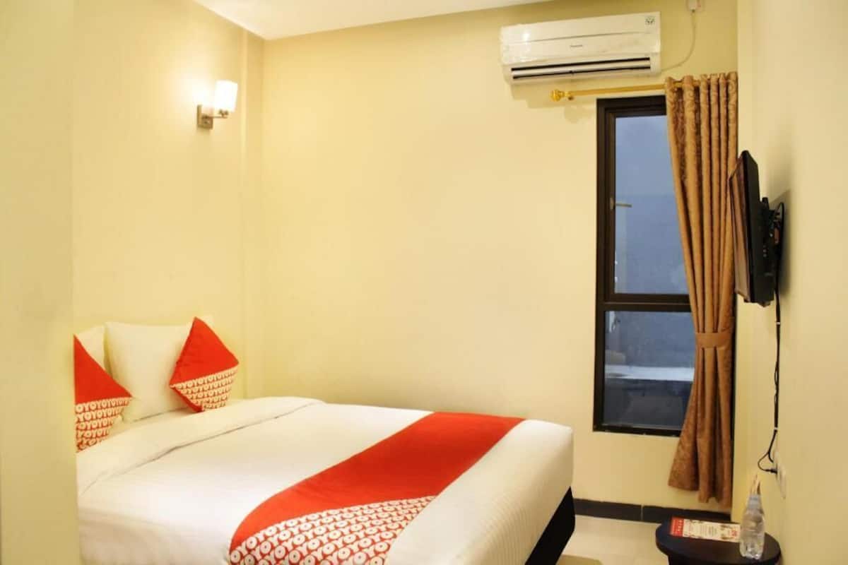 A hotel room with a bed, an air conditioner, a window with curtains, and a wall-mounted TV.