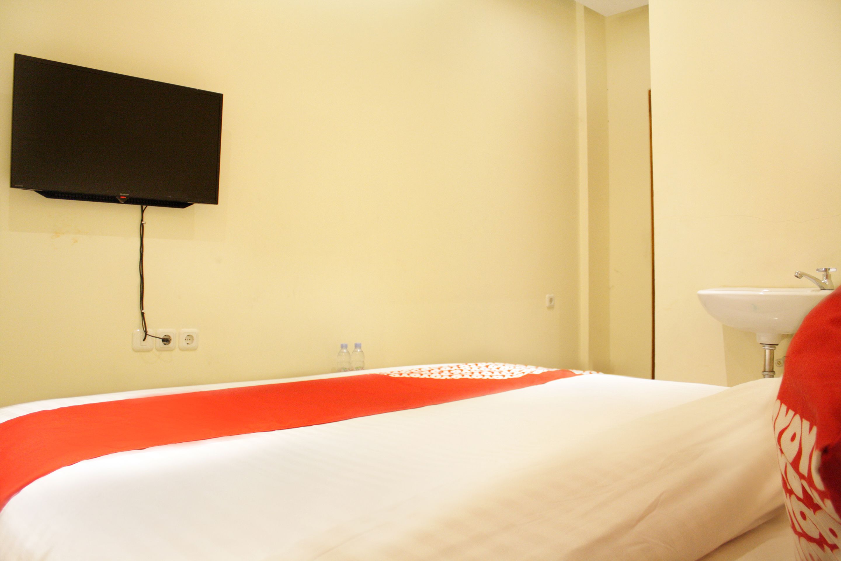 Habitación de hotel con cama, televisor montado, lavamanos y dos botellas sobre la mesita de noche.