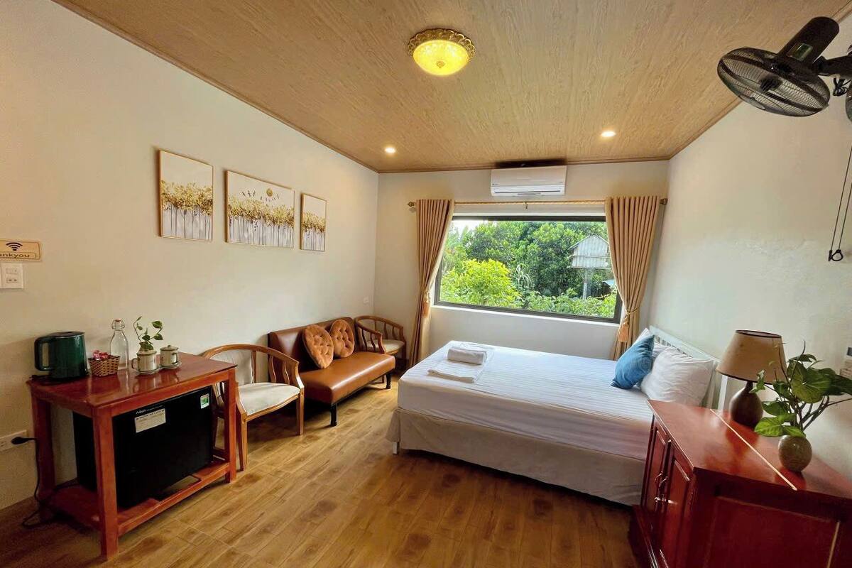 A room with a bed, a wooden table, a chair, a sofa, and a window with a view of greenery.