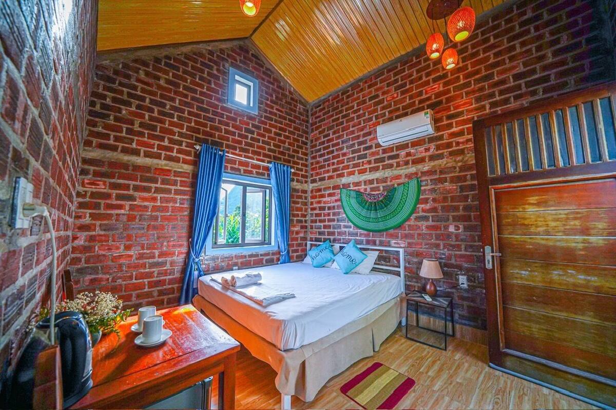 A room with a brick wall, a bed with pillows, a window with curtains, a table with a lamp, and a door.