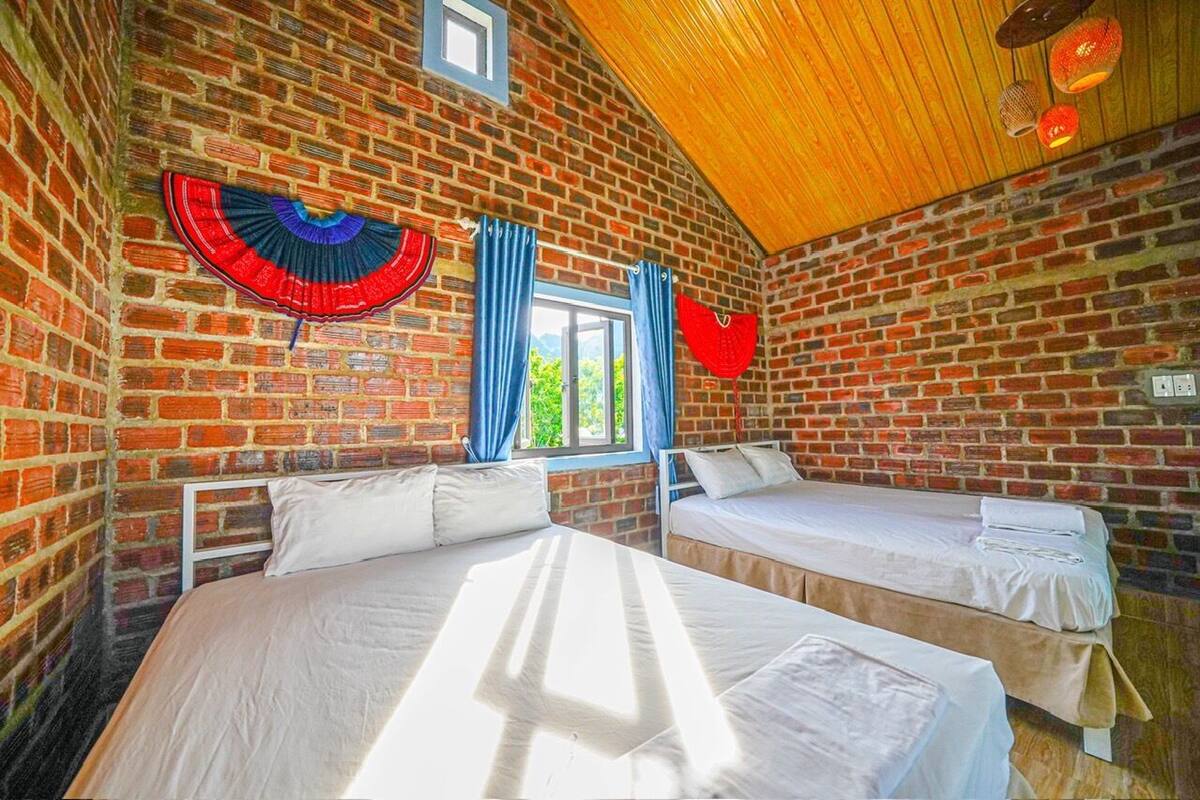 A room with two beds, brick walls, a wooden ceiling, and a window with blue curtains.
