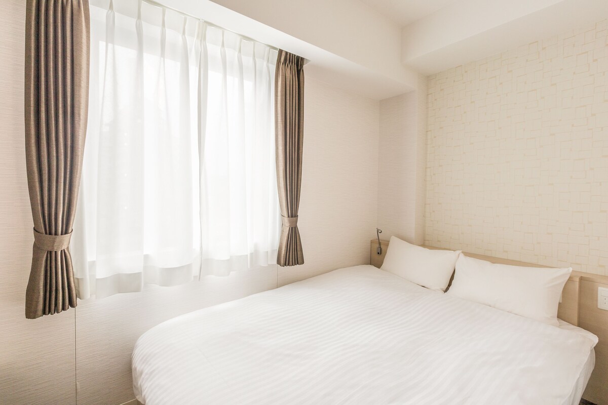 A neatly made bed in a hotel room with a plain wall and a window with curtains.