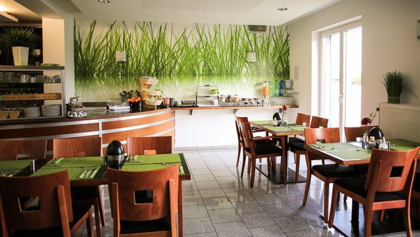 Un restaurant moderne avec des tables et des chaises en bois, un espace cuisine agrémenté d’un mur végétal, et un intérieur lumineux.