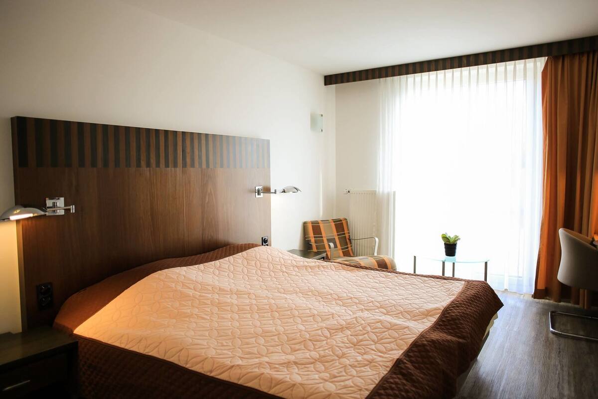 A hotel room with a large bed, a desk with a lamp, a chair, a small table with a plant, and a window with curtains.