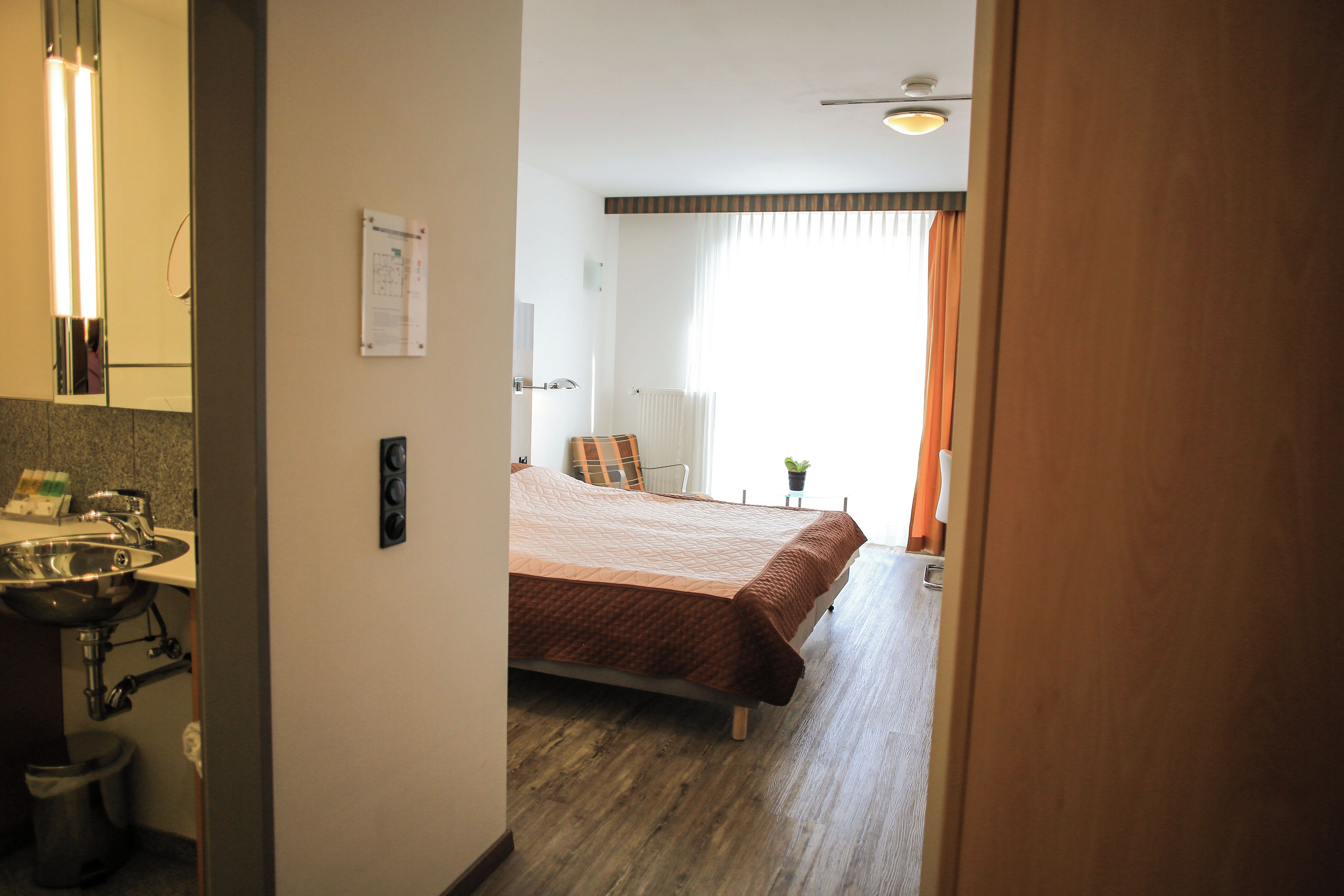 Ein Hotelzimmer mit Bett, Nachttisch, einem Fenster mit Vorhängen, einem Badezimmer mit Waschbecken und einem Spiegel.