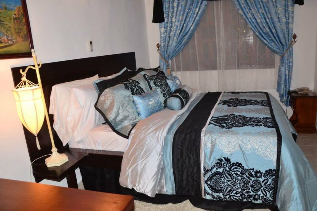 A neatly made bed with patterned bedding, a bedside lamp, and a window with blue curtains.