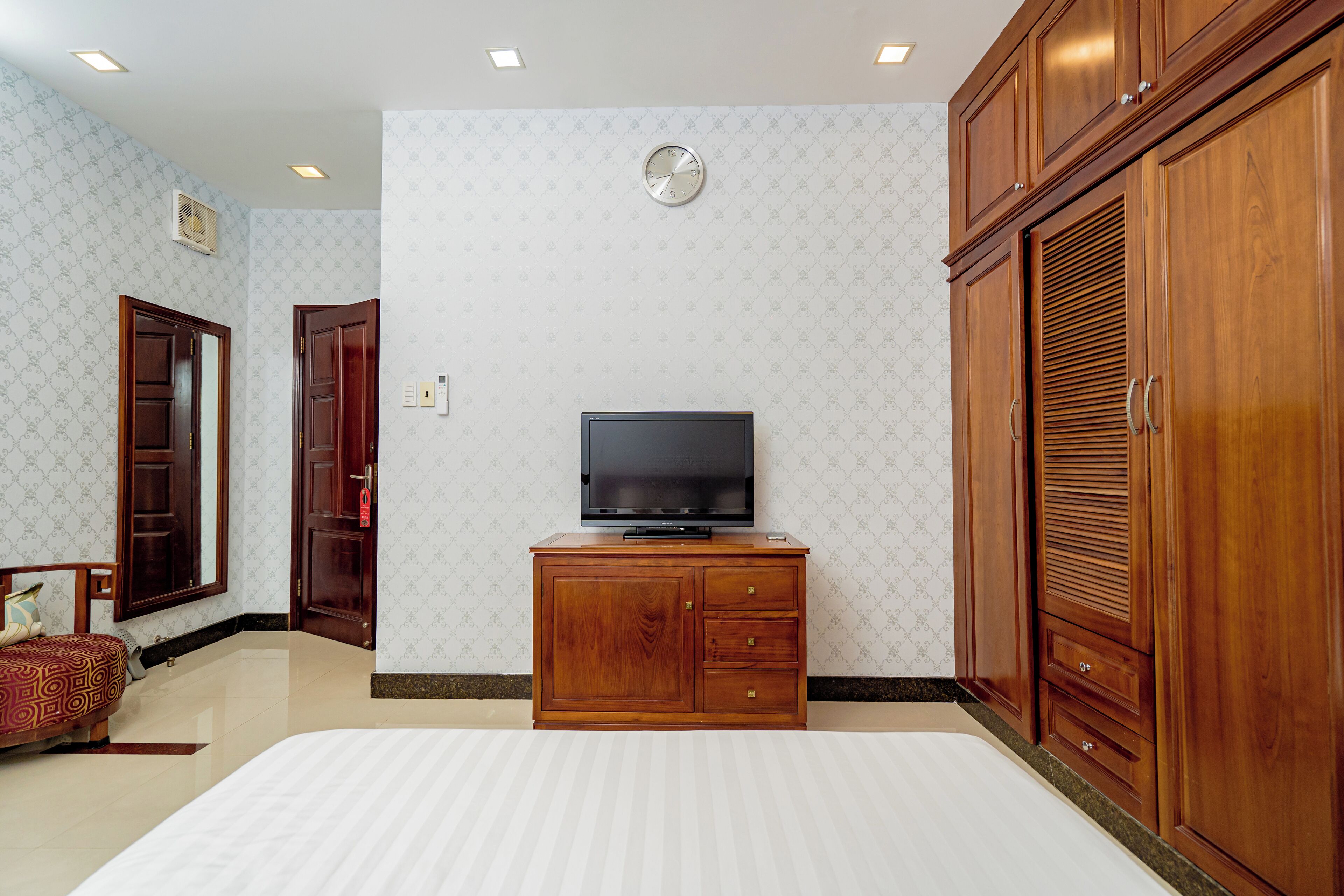 Habitación de hotel con cama, televisor sobre un mueble de madera, reloj en la pared y armarios de madera.