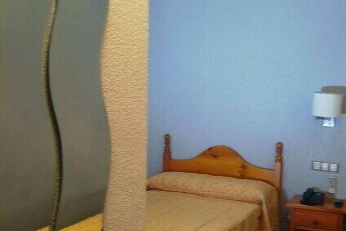 A single bed with a wooden headboard in a room with a plain wall and a nightstand with a lamp.