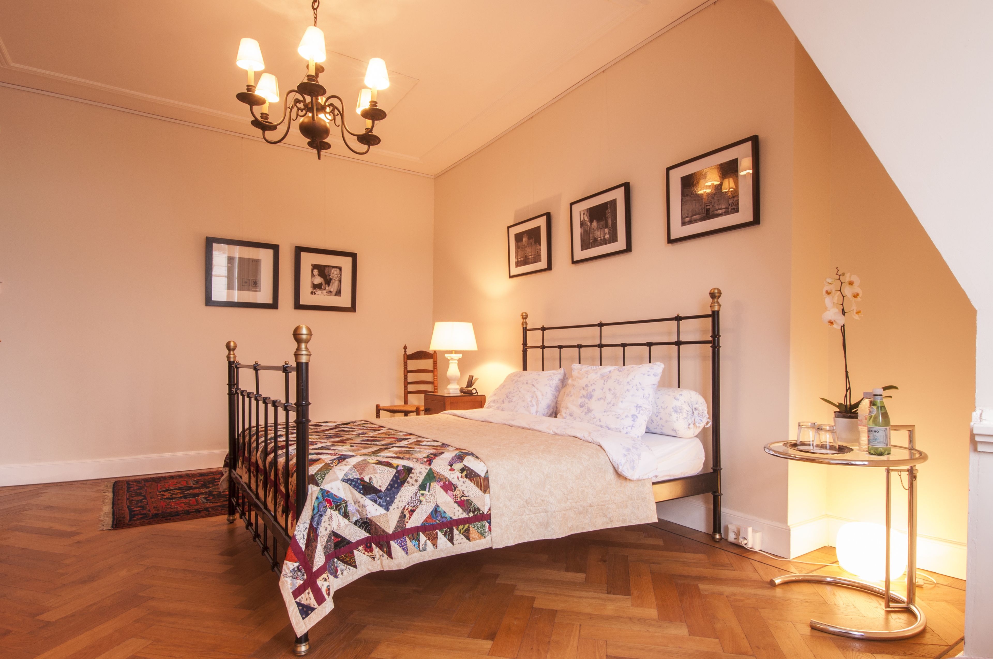 A bedroom with a bed, a chandelier, framed pictures on the wall, and a small table with a plant and a bottle.
