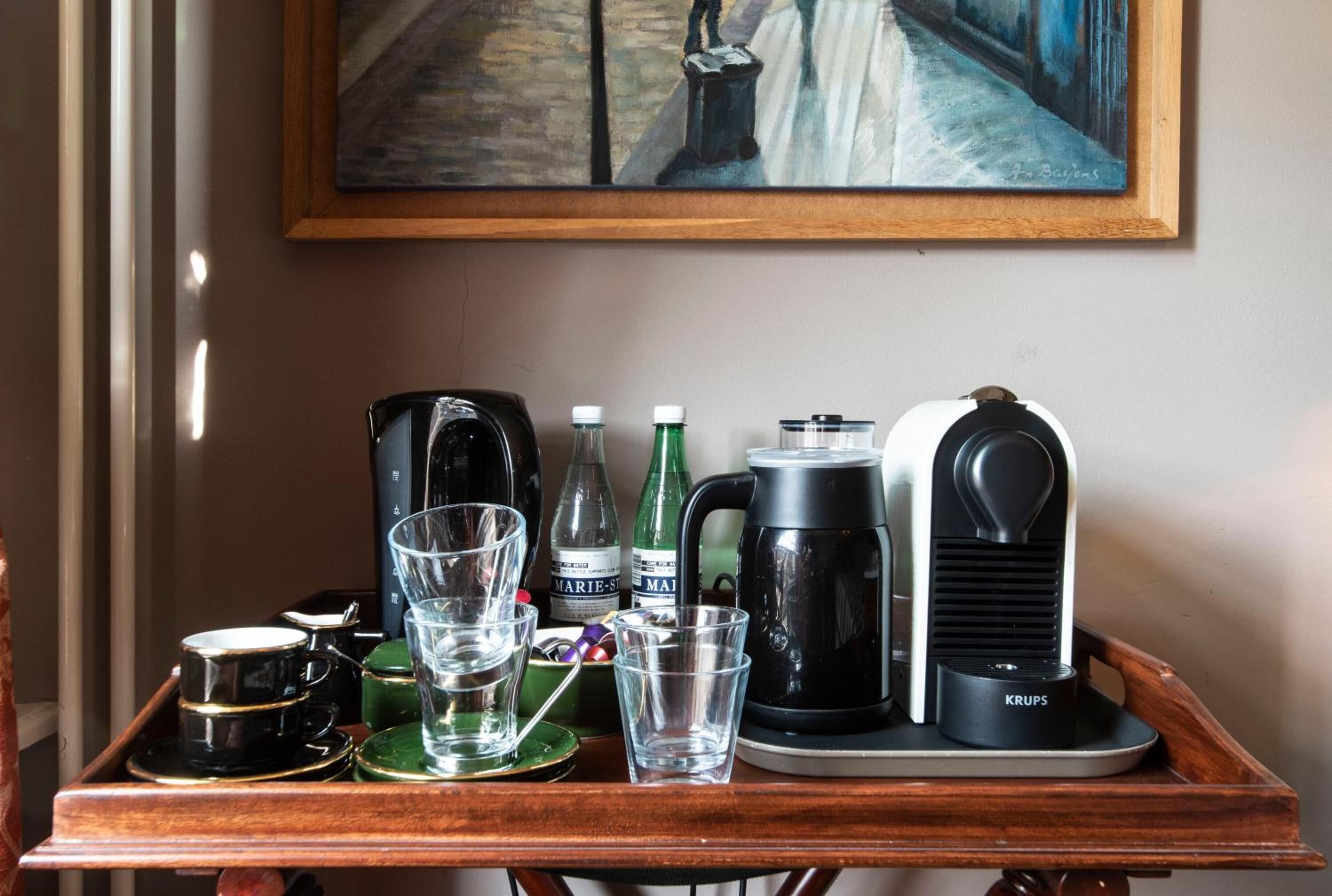 A wooden tray with a coffee maker, cups, glasses, and bottles on a table.