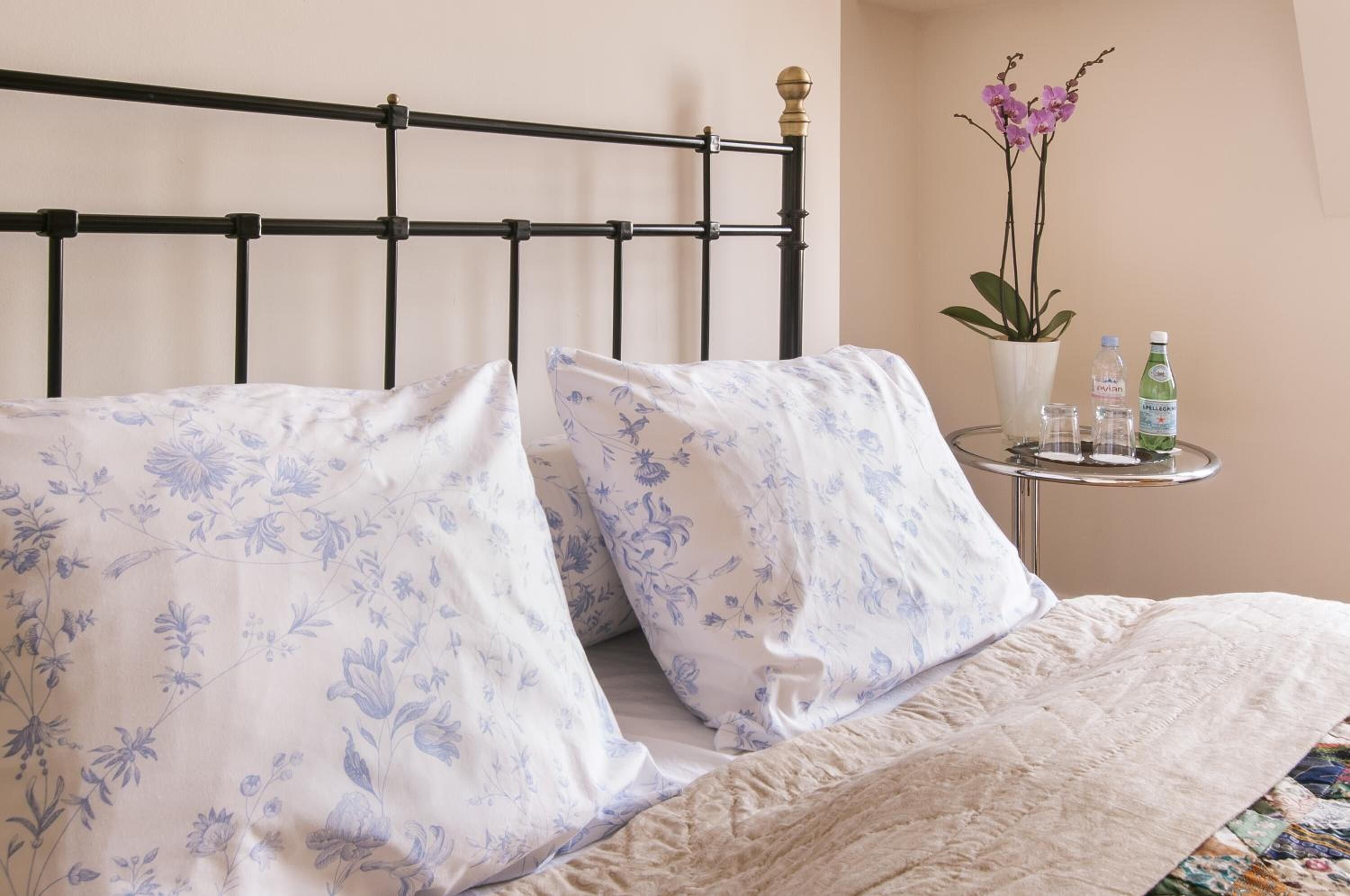 A bed with floral-patterned pillows, a round side table with a potted plant and bottles, and a simple metal headboard.