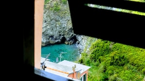 Vista costera a través de una ventana con persianas horizontales, mostrando un acantilado rocoso, agua turquesa y vegetación.