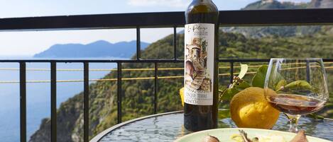 Una botella de vino, una copa de vino y un plato de comida en un balcón con vistas panorámicas del mar y las montañas.