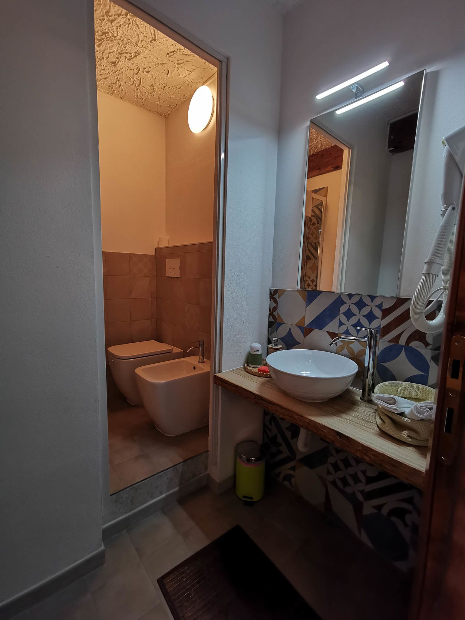 Une salle de bain moderne équipée de toilettes, d’un bidet et d’un lavabo. Un miroir est placé au-dessus du lavabo, et un porte-serviettes est fixé au mur. Les murs sont recouverts de carreaux disposés selon un motif géométrique.