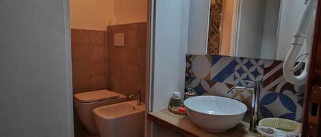 Une salle de bain moderne équipée de toilettes, d’un bidet et d’un lavabo. Un miroir est placé au-dessus du lavabo, et un porte-serviettes est fixé au mur. Les murs sont recouverts de carreaux disposés selon un motif géométrique.
