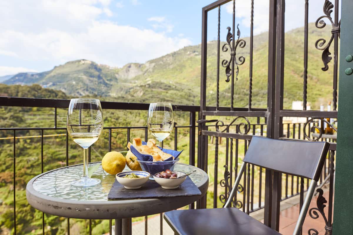 A balcony with a table set for two, featuring a glass of white wine, a bowl of olives, a lemon, and bread.