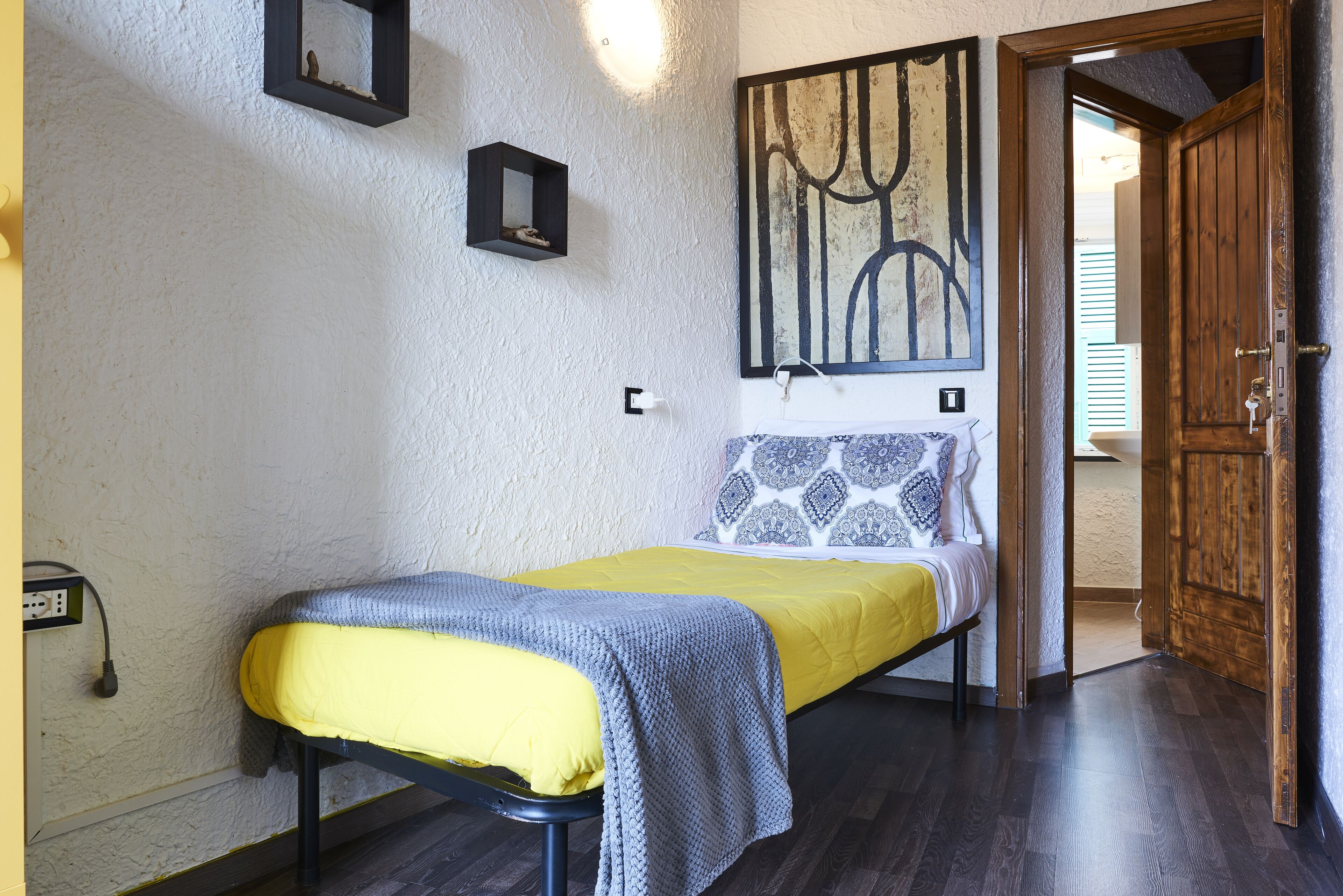 A single bed with a yellow and gray bedspread, a small bedside table, a wall-mounted light, and a door leading to another room.