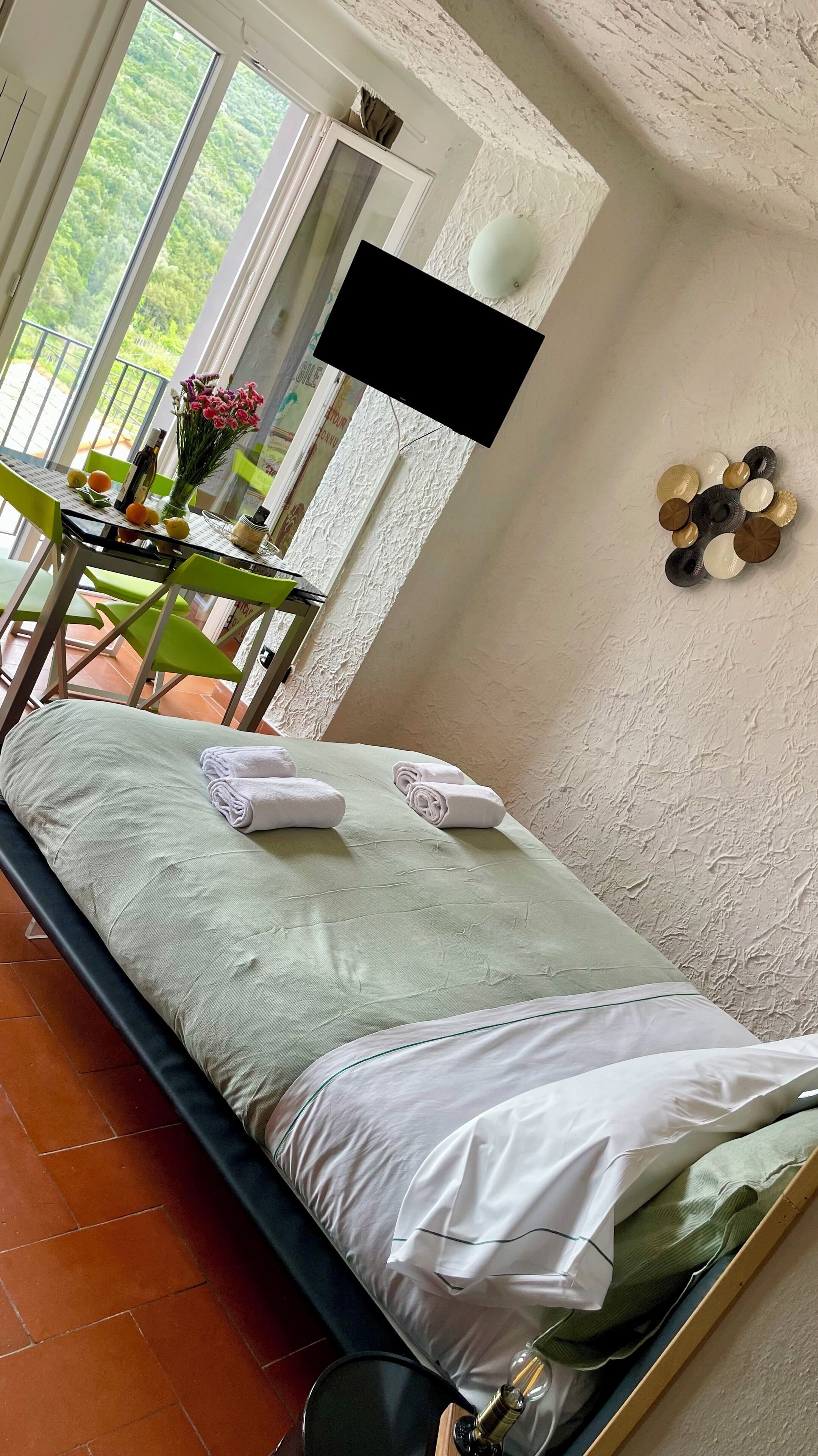 Una cama bien hecha con sábanas y toallas blancas, una mesa con una botella y flores, y vista a un espacio verde por la ventana.