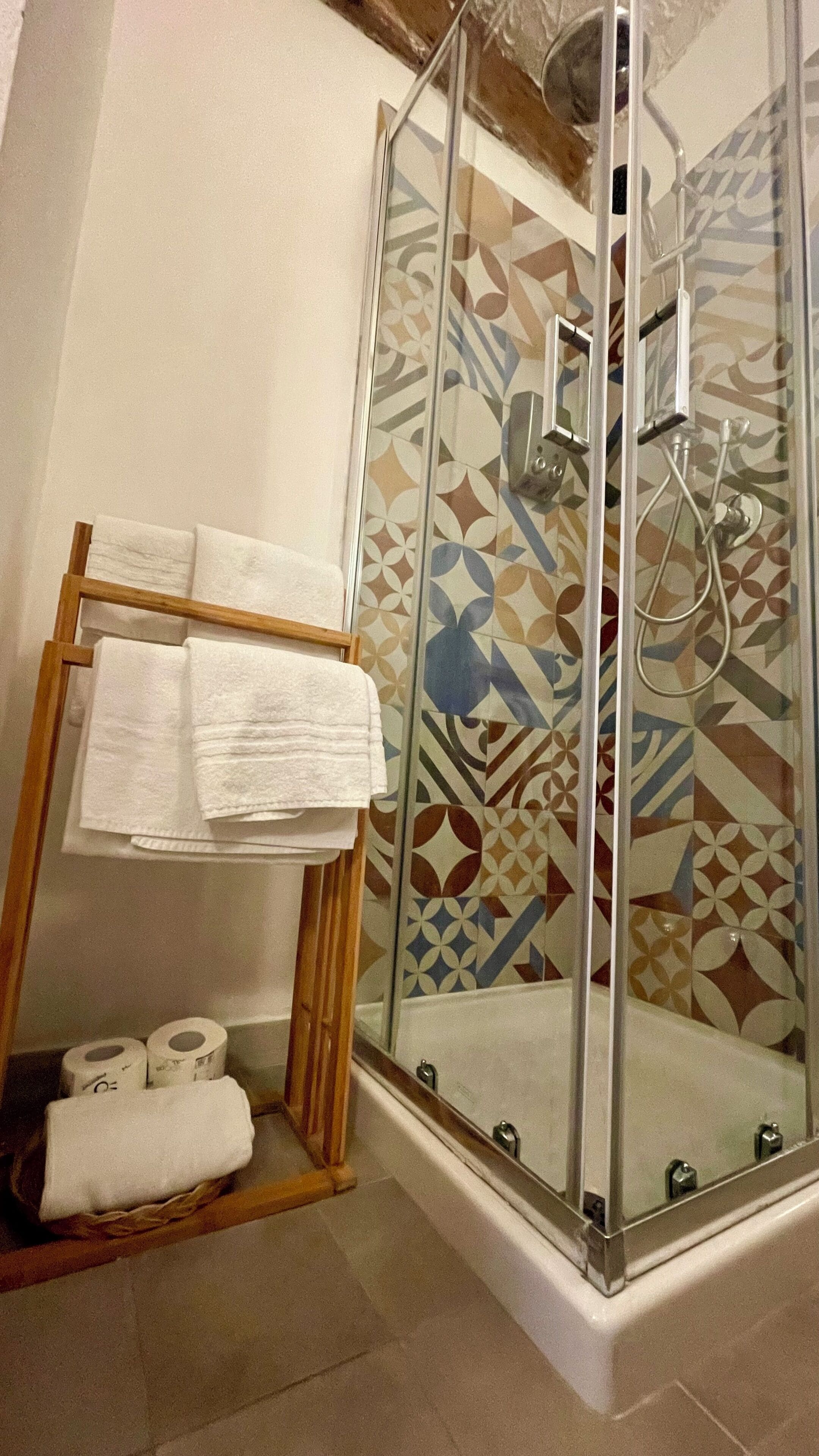 A glass-enclosed shower with a patterned tile wall, a wooden towel rack with folded towels, and two rolls of toilet paper on the floor.