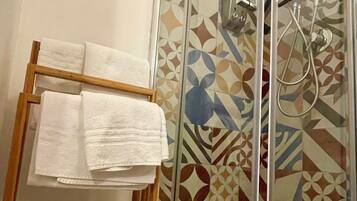 A glass-enclosed shower with a patterned tile wall, a wooden towel rack with folded towels, and two rolls of toilet paper on the floor.