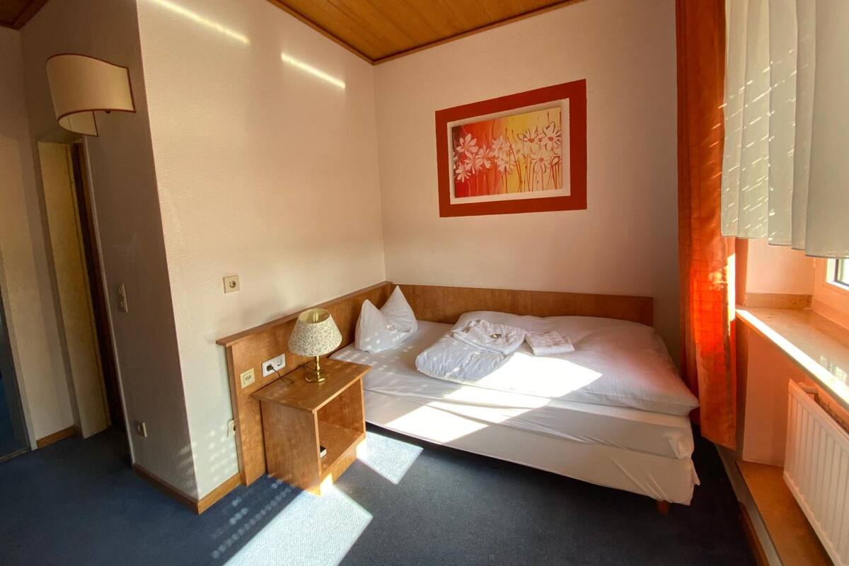 Un dormitorio con cabecera de madera, una mesita de noche con lámpara, cama con ropa blanca y un cuadro en la pared, además de una ventana con cortinas.