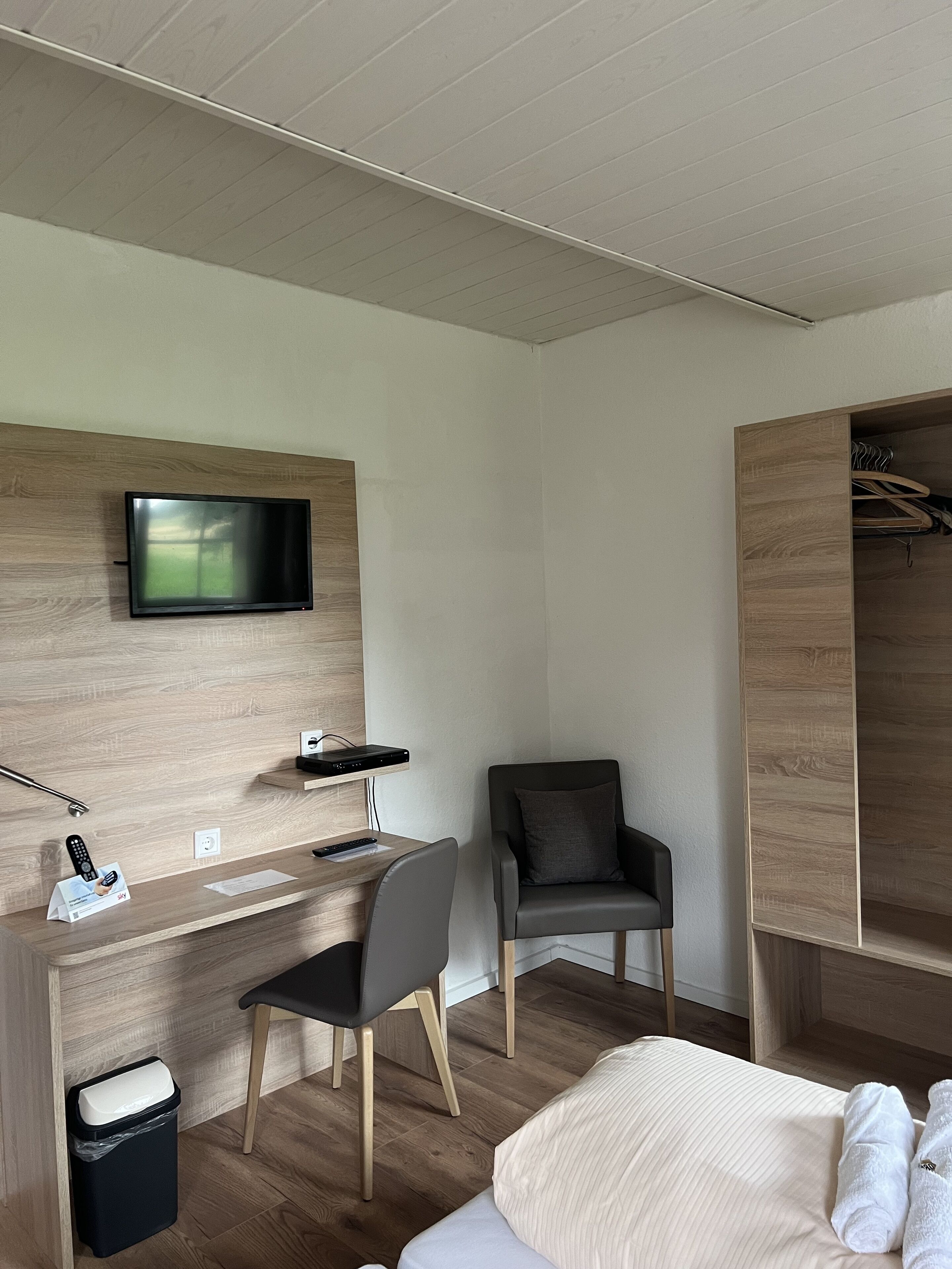 Una habitación de hotel moderna con un escritorio de madera, un televisor montado en la pared, una silla cómoda, una cama con sábanas blancas y un armario con espacio para colgar.
