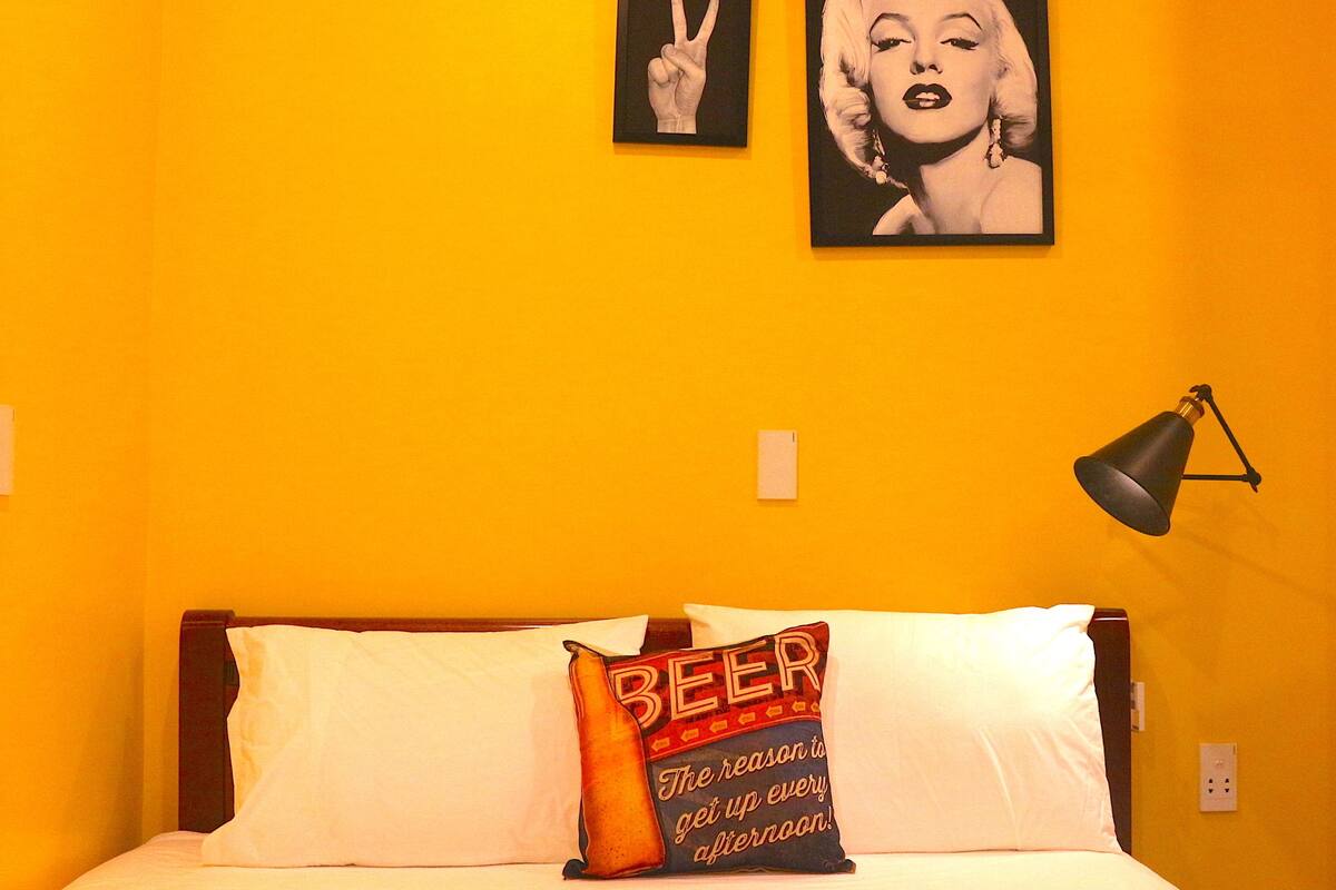 A hotel room with a bed, a pillow with 'BEER' text, a framed picture of a hand making a peace sign, and a lamp on the wall.