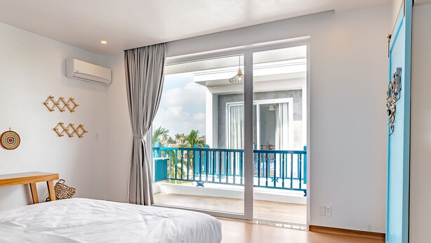 A bedroom with a bed, a wooden bench, a balcony with blue railings, and a view of palm trees. - The Present Home Hoi An (Da Nang)