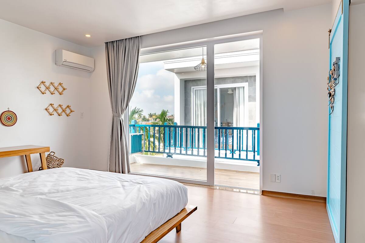 A bedroom with a bed, a wooden bench, a balcony with blue railings, and a view of palm trees.