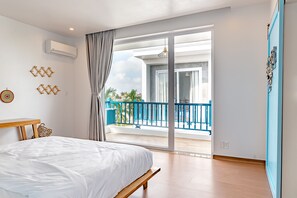 Un dormitorio con cama, banca de madera, balcón con barandillas azules y vista a palmeras.