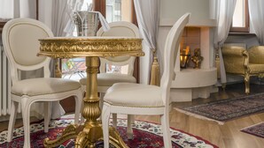 A room with a white and gold ornate table, two white upholstered chairs, a patterned rug, and a decorative fireplace.