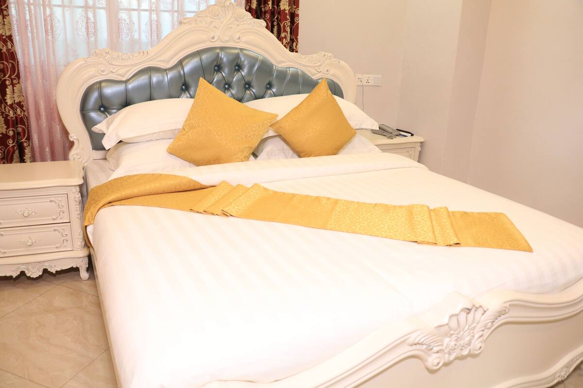 A neatly made bed with a white and gold color scheme, a decorative headboard, and a bedside table with a drawer.