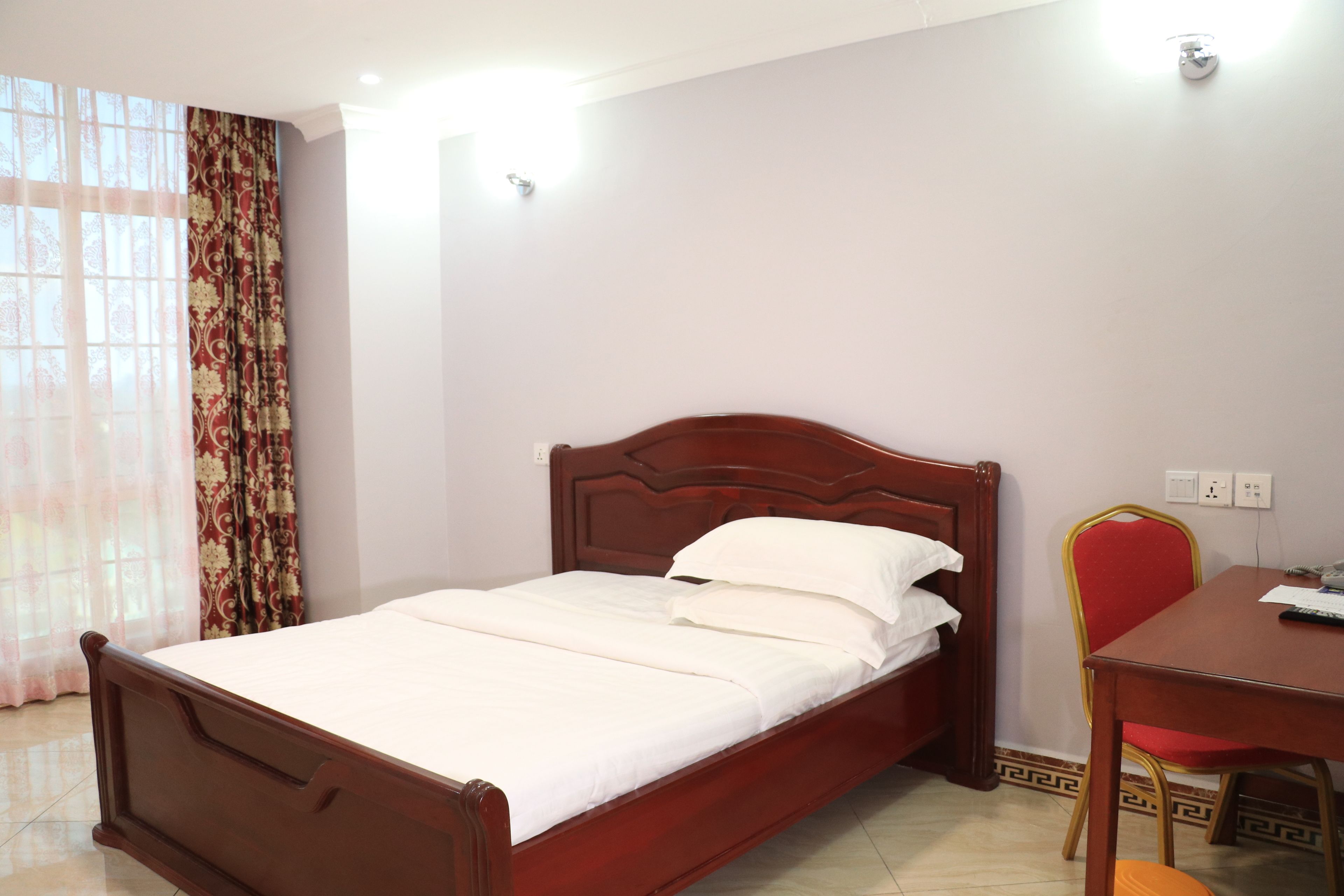 Uma cama bem arrumada com cabeceira de madeira, cortina estampada em vermelho e dourado, e uma pequena mesa com uma cadeira ao lado.
