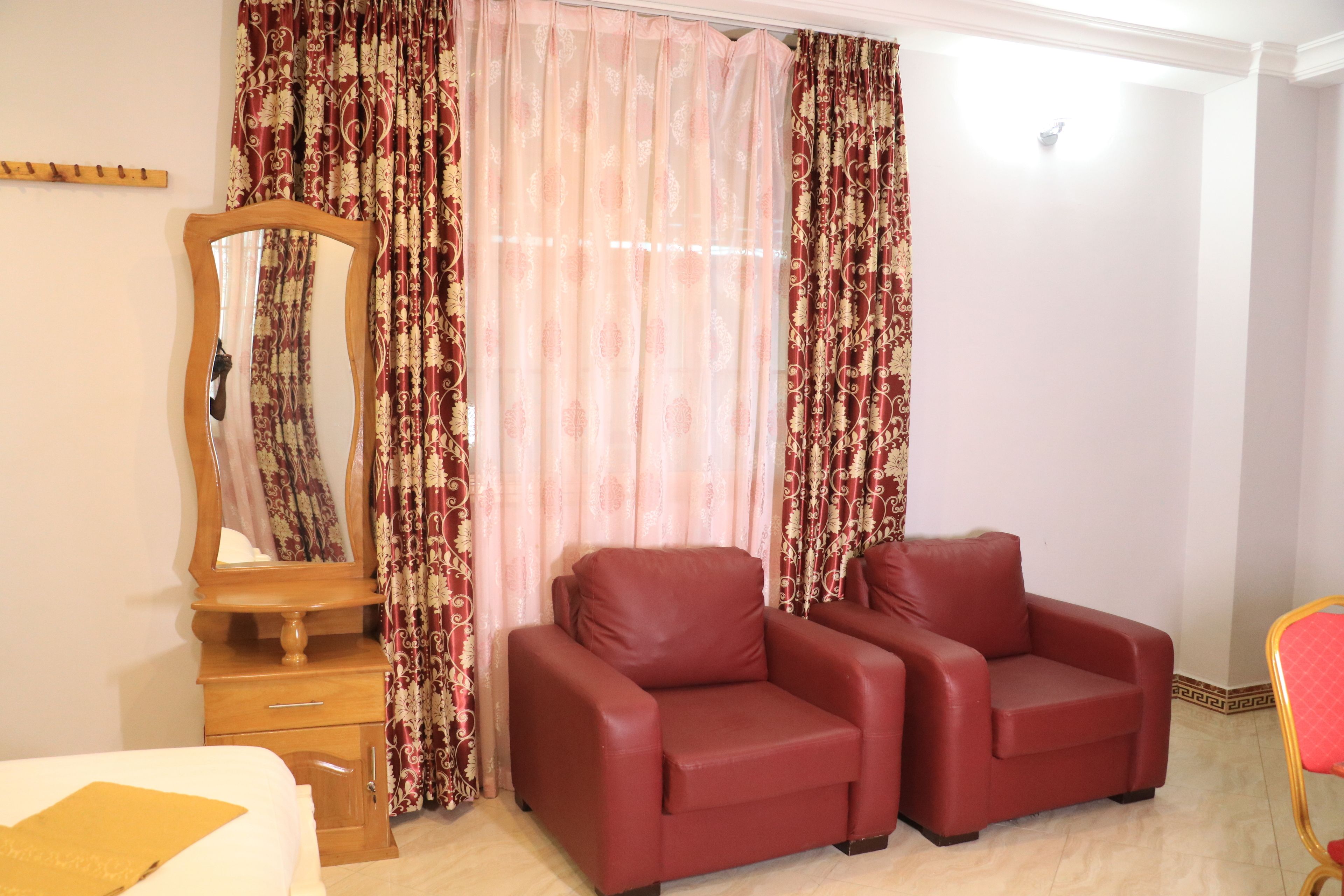 Una habitación con una cama, un aparador de madera, un espejo, dos sillones de cuero rojo y cortinas estampadas.