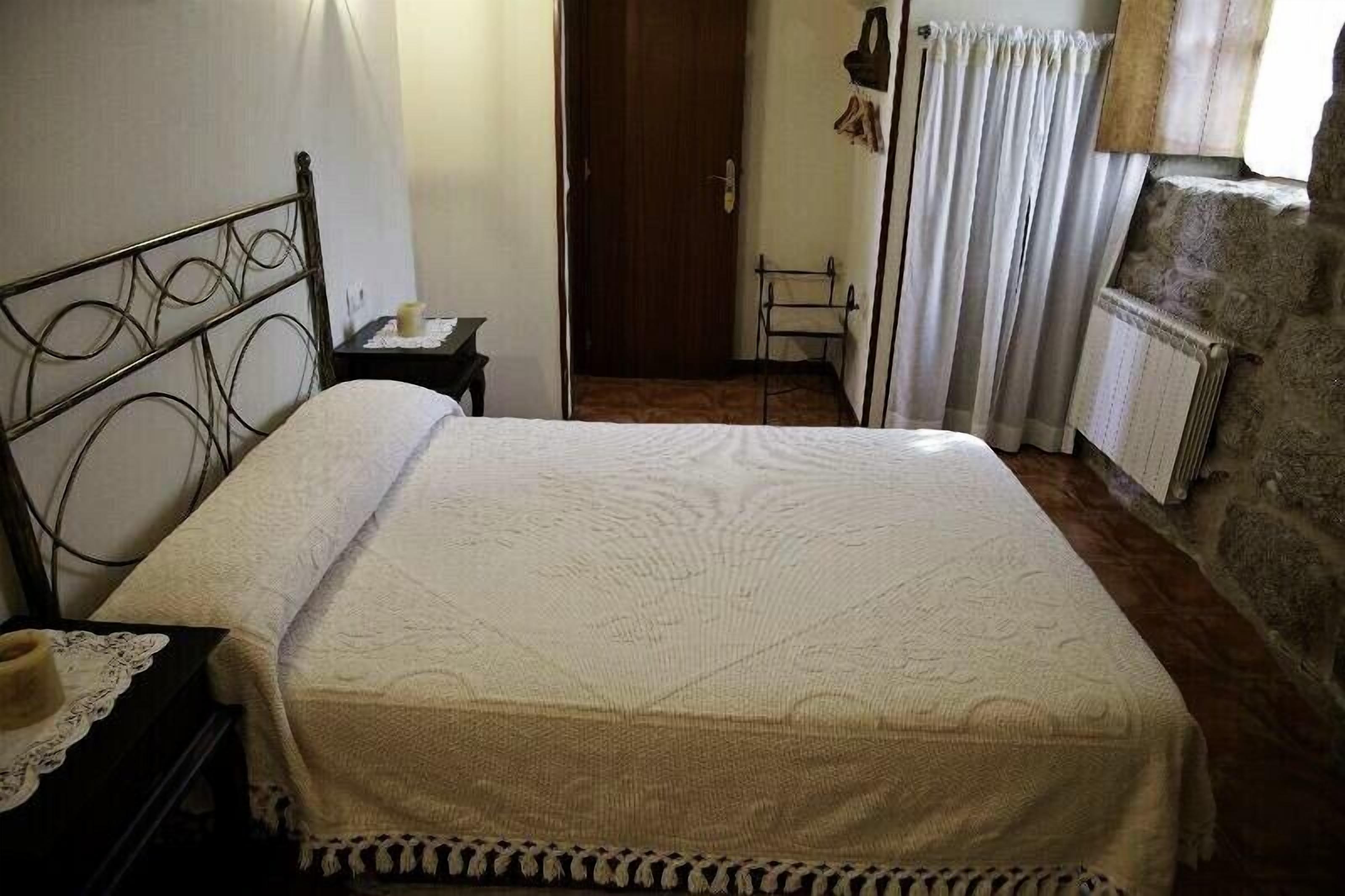 Un dormitorio con estructura de cama metálica, colcha blanca, mesita de noche con lámpara, una puerta que da a otra habitación y pared de piedra.