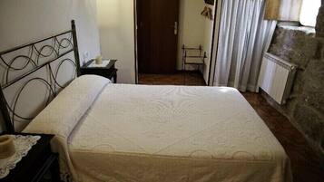 Un dormitorio con estructura de cama metálica, colcha blanca, mesita de noche con lámpara, una puerta que da a otra habitación y pared de piedra.