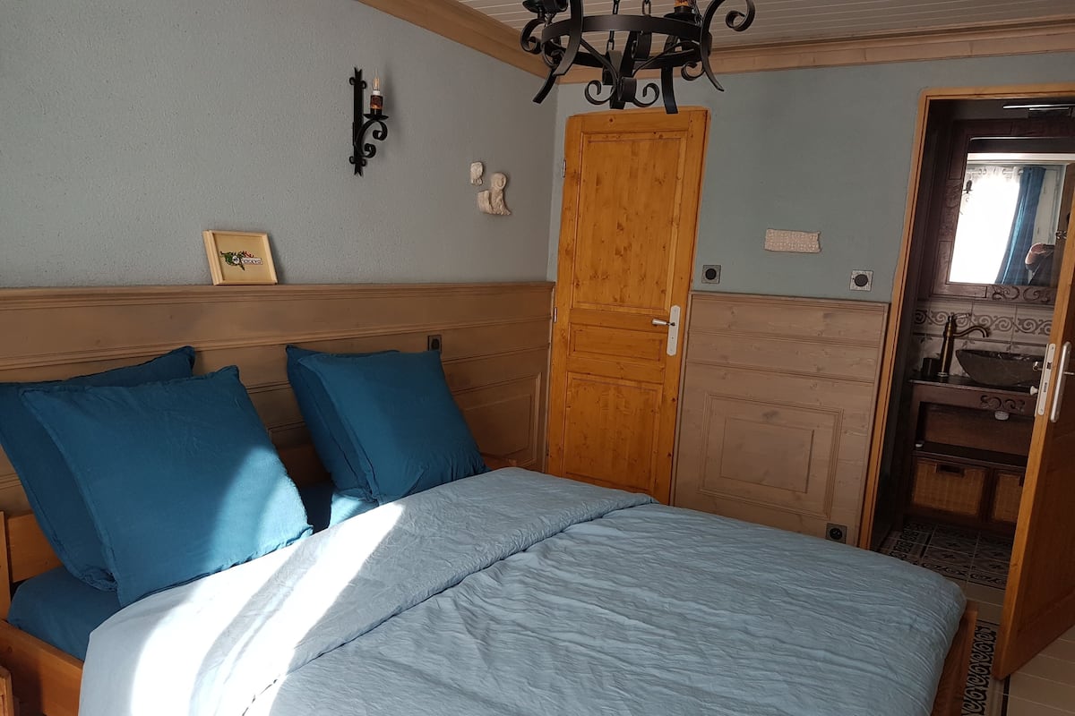 Une chambre à coucher avec une tête de lit en bois, un lit avec une literie bleue, une porte en bois, un luminaire fixé au mur et une petite étagère avec un tableau.