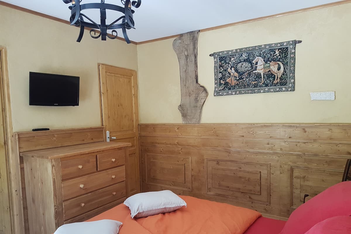 Une chambre à coucher avec une tête de lit en bois, un téléviseur fixé au mur, une tapisserie décorative et un lit avec une literie et des oreillers orange.