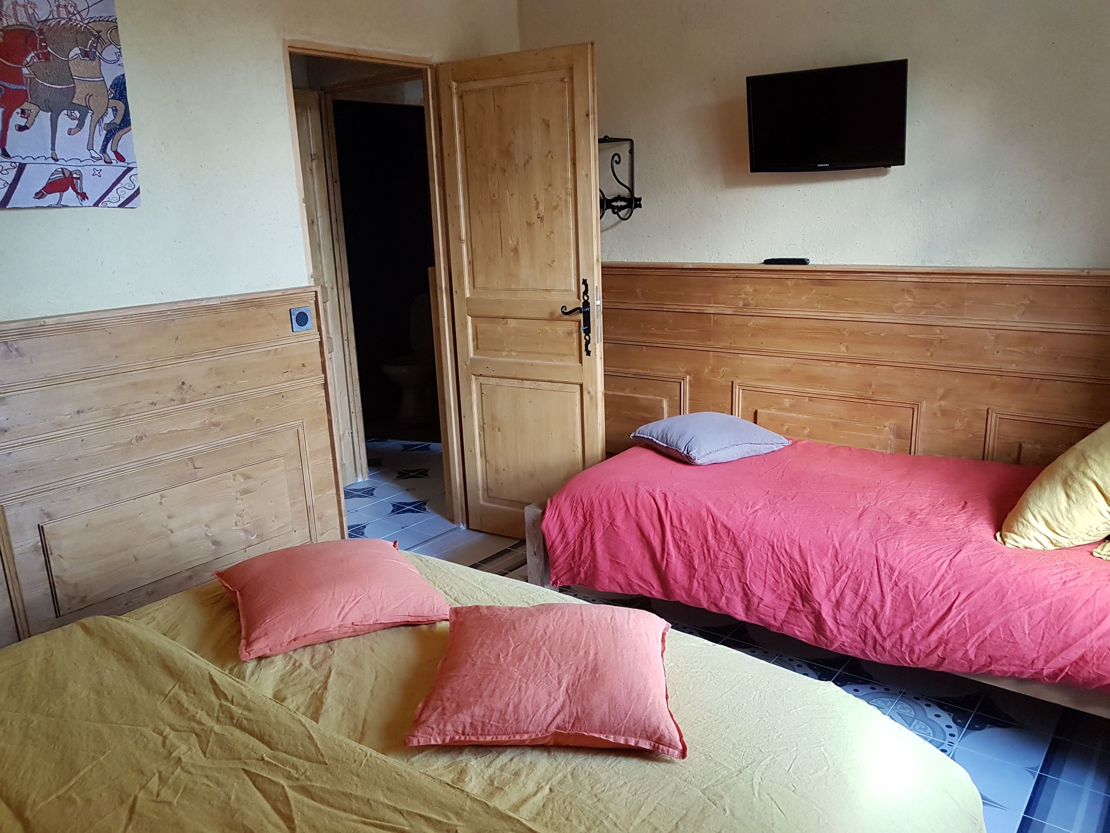 A bedroom with two beds, one with pink bedding and the other with yellow bedding. There is a wooden headboard, a wall-mounted TV, and a door leading to another room.