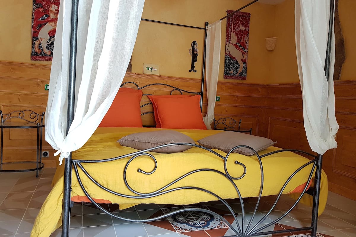 Un lit à baldaquin avec une couvre-lit jaune et des oreillers orange, une tête de lit en métal et un mur en bois orné d’une tapisserie.
