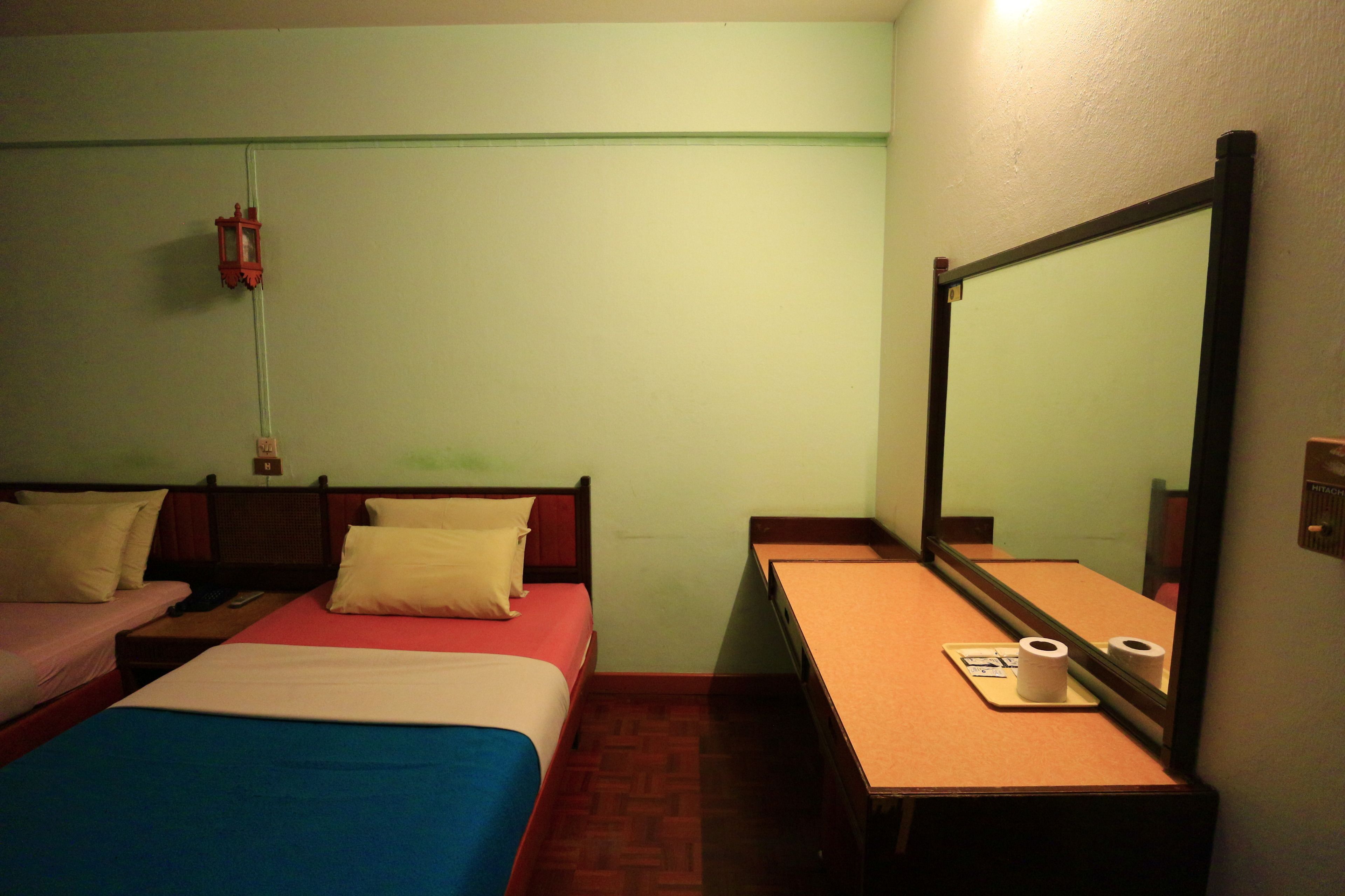 A small hotel room with two beds, a desk, and a mirror. There is a wall socket and a fire alarm.