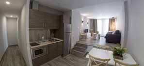 A modern kitchen with wooden cabinets, a built-in oven, and a refrigerator. There is a dining area with a white table and chairs, and a living room with a sofa and a TV. - Hotel Asador O Callejón De Belchite (Ayerbe)