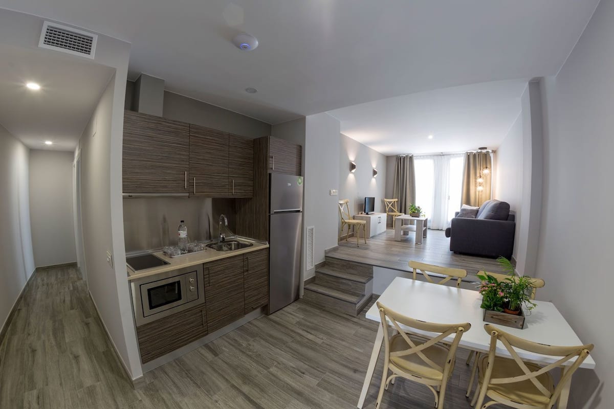 Un appartement moderne comprenant une cuisine, un coin repas et un salon. Il est doté d’un plancher en bois, d’un four encastré, d’un réfrigérateur et d’une table à manger avec des chaises.