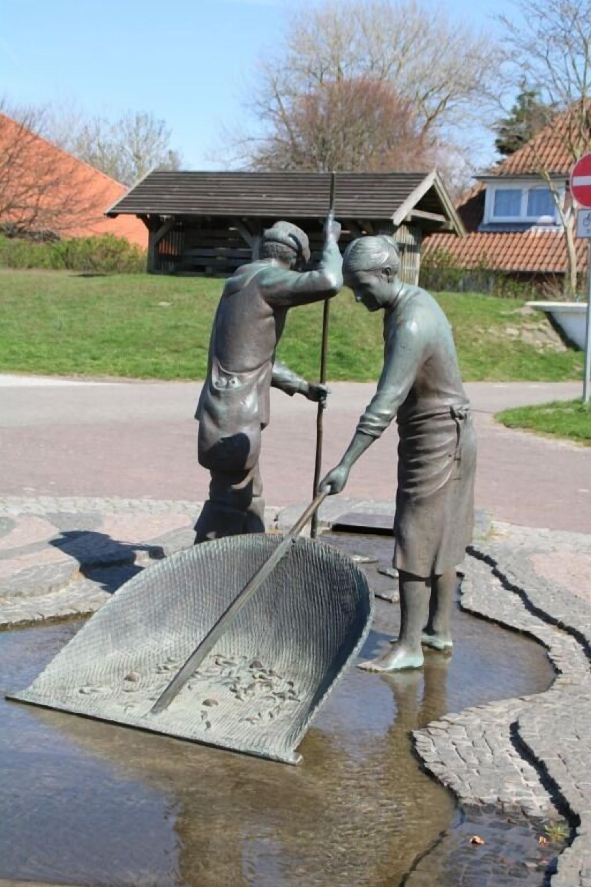 Eine Statue von zwei Figuren, eine mit Schaufel und die andere mit Rechen, in einem öffentlichen Platz mit gepflastertem Bereich und einem Gebäude im Hintergrund.