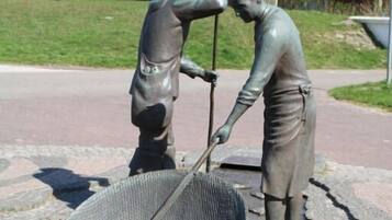 Eine Statue von zwei Figuren, eine mit Schaufel und die andere mit Rechen, in einem öffentlichen Platz mit gepflastertem Bereich und einem Gebäude im Hintergrund.