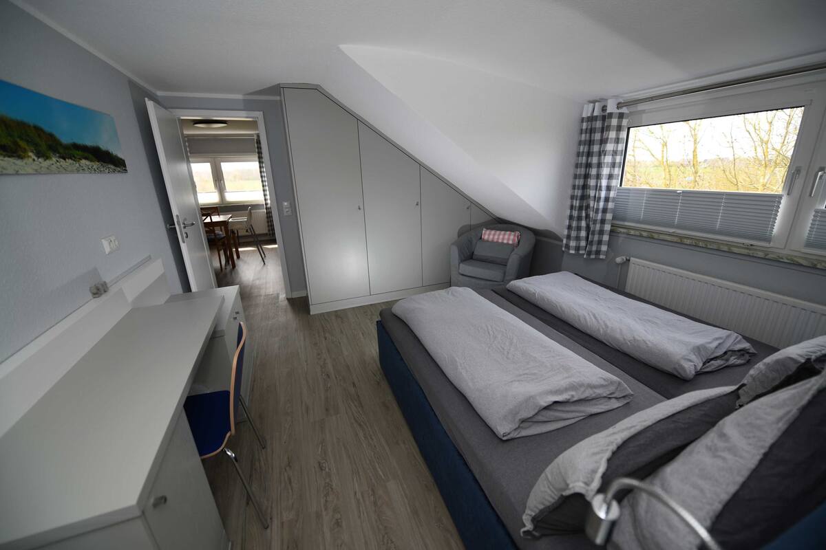 A compact bedroom with a bed, a desk, and a chair. There is a wardrobe and a window with curtains.