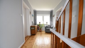 A hallway with wooden flooring, a staircase with wooden railings, a grey sofa, a wooden cabinet, and a potted plant. - Landhaus Dircks (St. Peter-Ording)