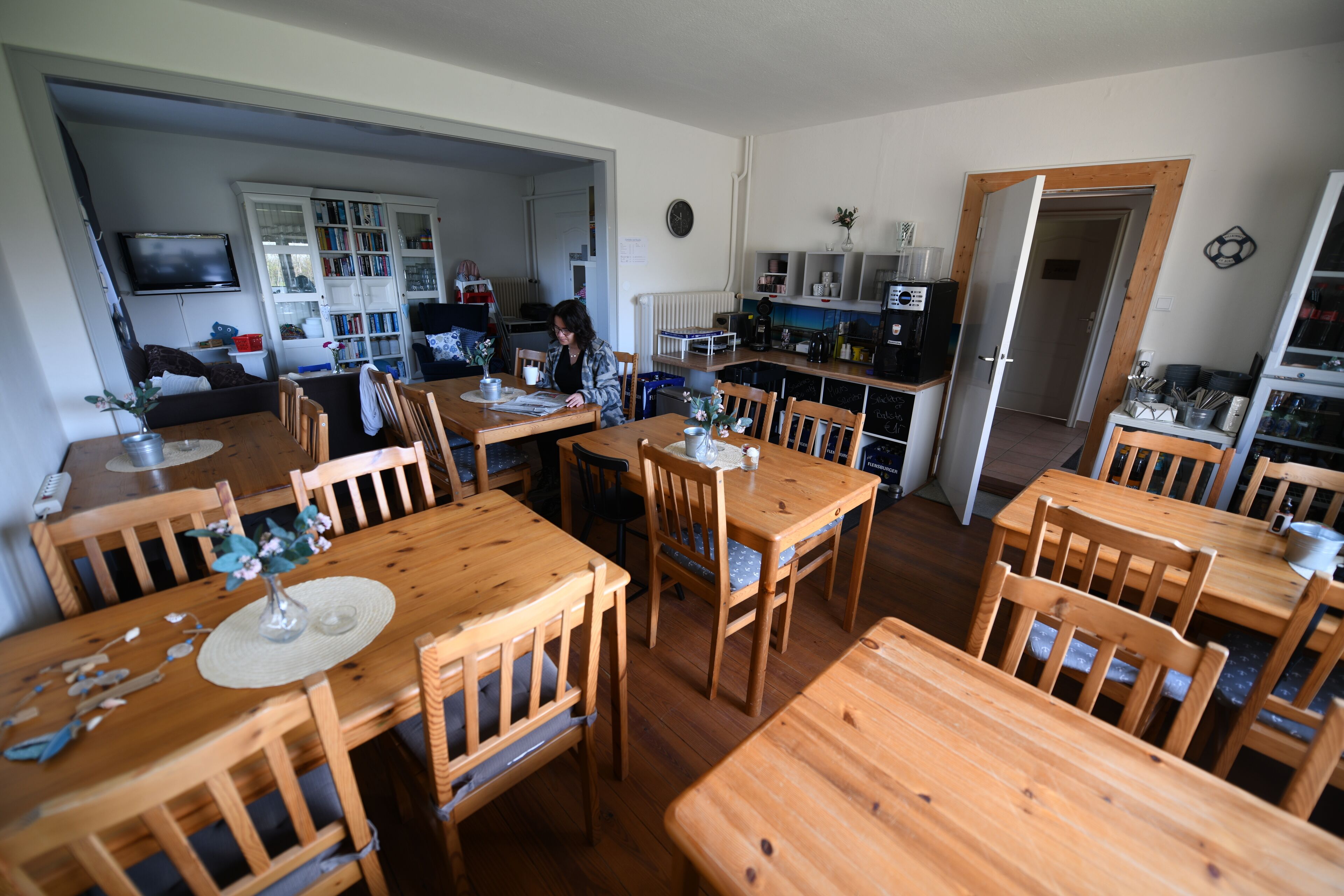 A dining area with wooden tables and chairs, a kitchen in the background, and a television mounted on the wall.